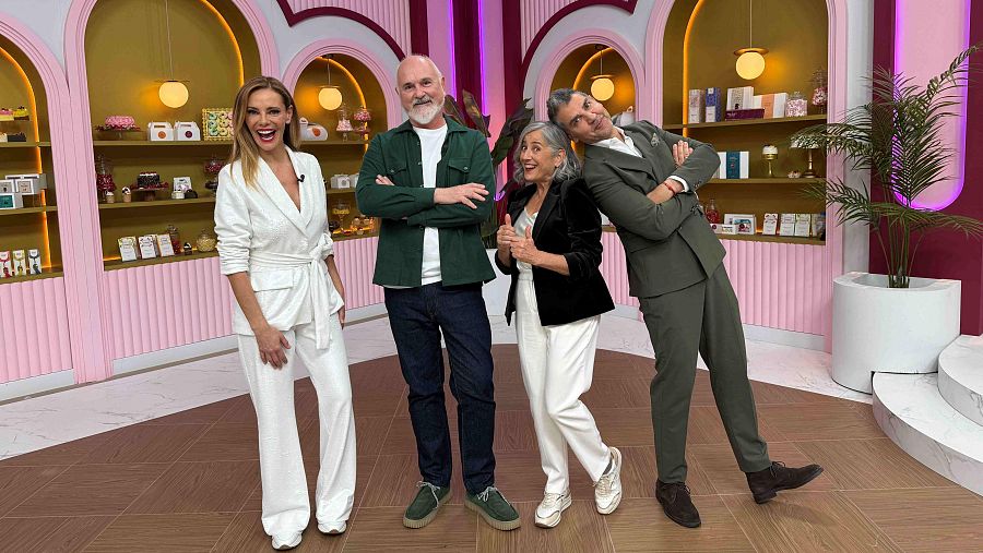 En un set televisivo con decoración de pastelería, cuatro individuos posan. Destacan una mujer de blanco sonriente, un hombre barbudo con camisa verde, una mujer con chaqueta negra y un hombre con traje verde oliva.