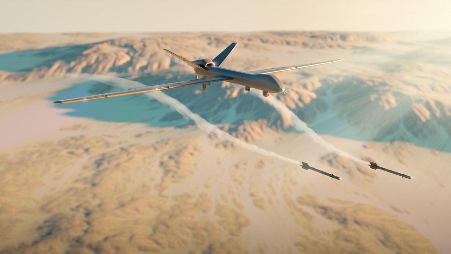 Un dron militar grisáceo, en un entorno desértico con montañas, dispara tres misiles que dejan una estela de humo blanco.