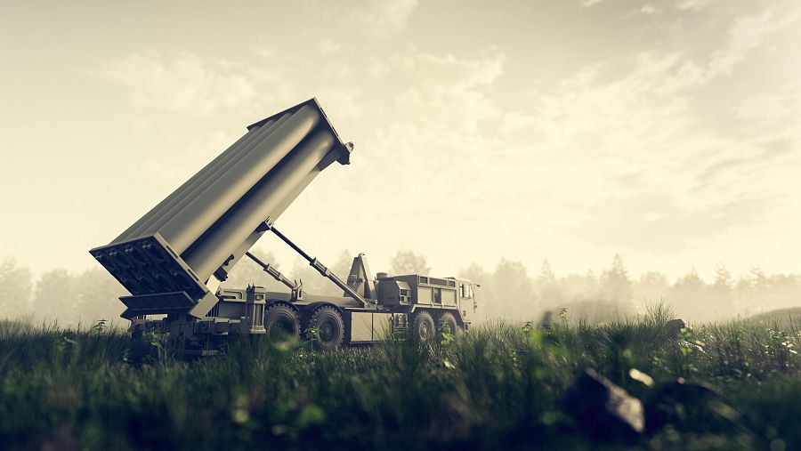 Un vehículo militar THAAD, con el lanzador elevado, se encuentra en un campo cubierto de hierba alta, con árboles difuminados en el horizonte y un cielo nublado.