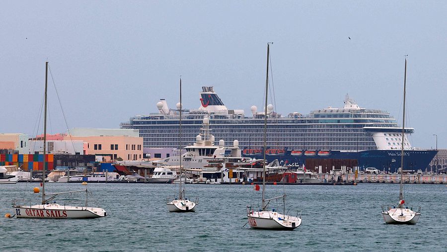 Un gran crucero azul oscuro, el "Mein Schiff 5", se encuentra anclado en el puerto, con veleros blancos en primer plano y edificios coloridos al fondo.
