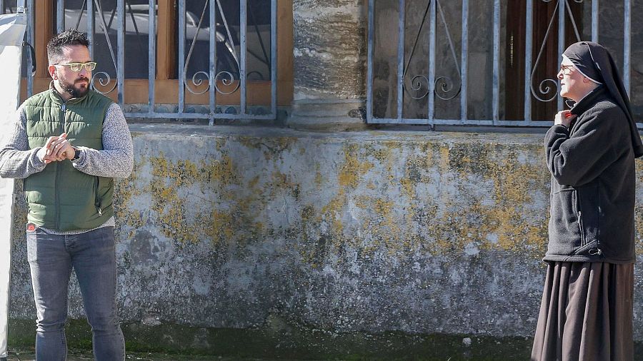 Un hombre barbudo con gafas y un chaleco verde, junto a una monja con hábito, posan frente a un edificio con ventanas enrejadas y una fachada desgastada.