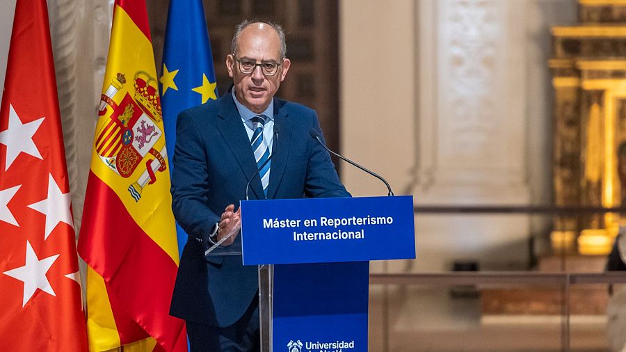 Un hombre con gafas y traje azul, posiblemente José Antonio Guardiola, se encuentra frente a un atril con el texto "Máster en Reporterismo Internacional" y el logo de una universidad, con banderas detrás.