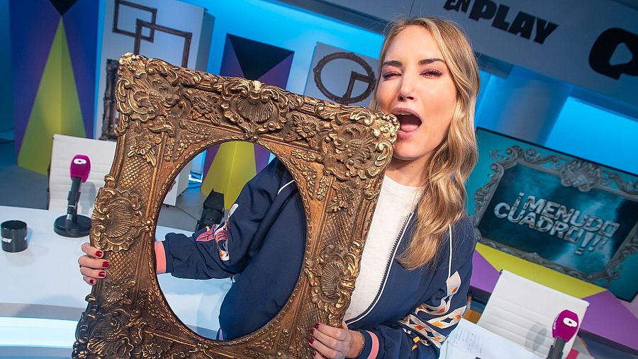 Alba Carrillo, con una chaqueta azul y uñas rojas, interactúa con un marco dorado, sacando la lengua. Un micrófono rosa y el logo de un programa televisivo son visibles en el entorno.