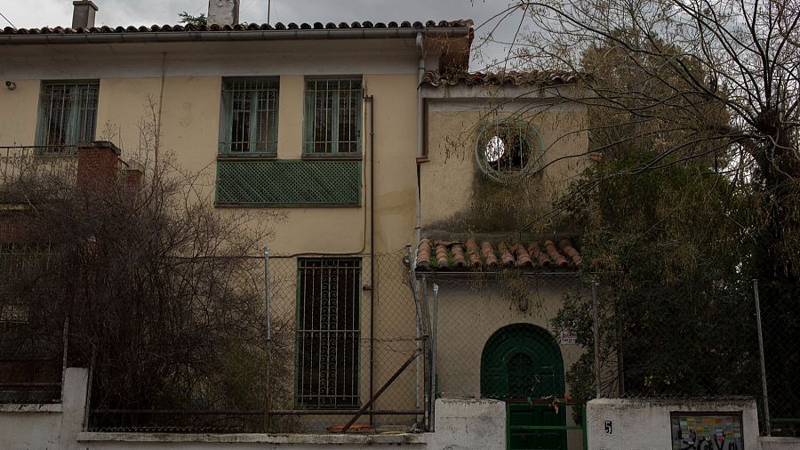 Fachada de una casa de dos plantas con ventanas y balcón con rejas verdes, puerta arqueada verde oscuro y tejado de tejas rojas, rodeada por una valla metálica y vegetación.