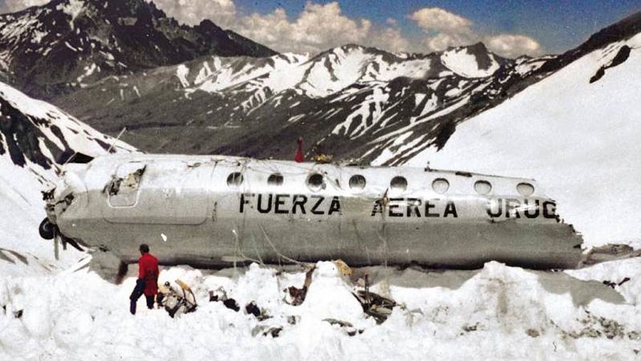 Un avión dañado con la inscripción "FUERZA AEREA URUG" yace en un paisaje montañoso nevado. Una persona vestida con una chaqueta roja se encuentra cerca de los restos.