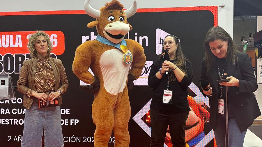 Un grupo de personas, incluyendo una disfrazada de toro con un medallón, posan para la foto. Una mujer con gafas y camiseta de RTVE sostiene un micrófono, mientras que otra revisa su teléfono móvil.