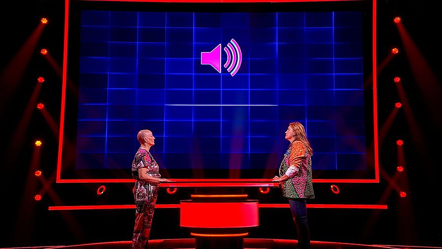 En un escenario con luces rojas, dos mujeres se preparan frente a una pantalla con un icono de altavoz. Una de ellas viste un conjunto estampado en tonos rojos, y la otra, una chaqueta con motivos geométricos y vaqueros.