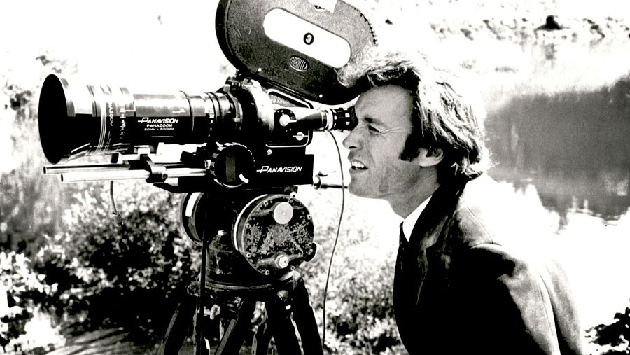 Clint Eastwood, en blanco y negro, mira a través de una cámara de cine montada en un trípode, con un paisaje natural de fondo.