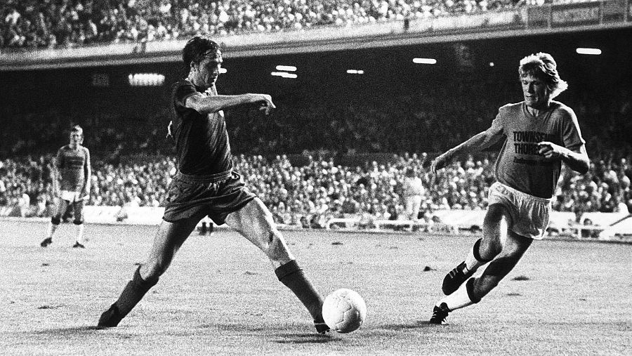 Imagen en blanco y negro de un partido de fútbol donde un jugador con camiseta oscura se dispone a golpear el balón, con otro jugador acercándose. El encuentro se desarrolla en un estadio lleno de gente.