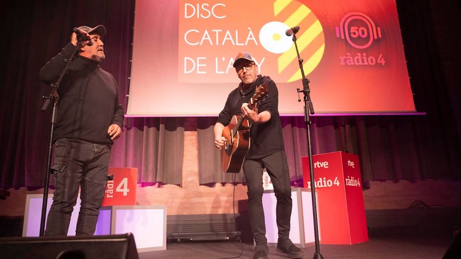 Una actuació en directe de Lax'n'Busto amb dos membres a l'escenari; un cantant amb gorra i un guitarrista amb gorra, amb una pantalla al fons amb el logo de Ràdio 4.