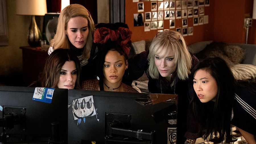 Un grupo de mujeres, incluyendo a Sandra Bullock, Sarah Paulson, Rihanna, Cate Blanchett y Awkwafina, miran fijamente dos pantallas de ordenador con expresiones de sorpresa. El entorno es informal, con fotos y pegatinas en la pared y en las pantallas.