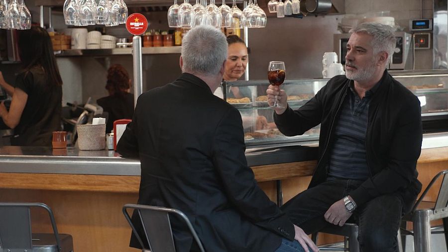 En una barra de bar, Adrià Collado, amb jaqueta negra i samarreta de ratlles, sosté una copa. Darrere, una altra persona i una empleada atenen el local, amb una màquina de cervesa Estrella Damm.