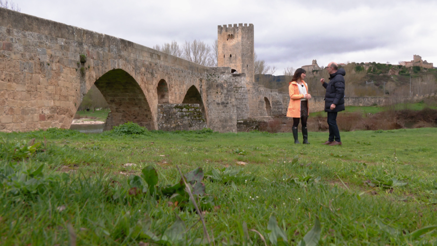 Dos personas dialogan frente a un puente de piedra con arcos y una torre central. La mujer lleva un abrigo naranja y falda oscura, y el hombre viste una chaqueta oscura y vaqueros. Un castillo se vislumbra en la distancia, sobre una colina.