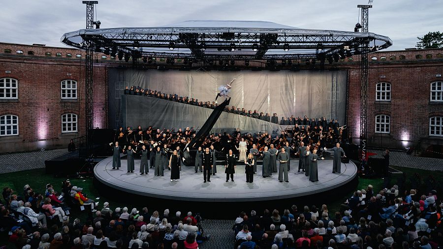 Espectáculo en la Fortaleza de Oscarsborg, Noruega, con orquesta, coro, solistas y artista aéreo. Edificios históricos iluminados y público atento.