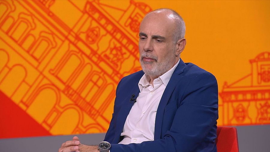 Joan Ridao, con camisa blanca y americana azul, posa en un plató con fondo naranja. Su expresión es seria y reflexiva.