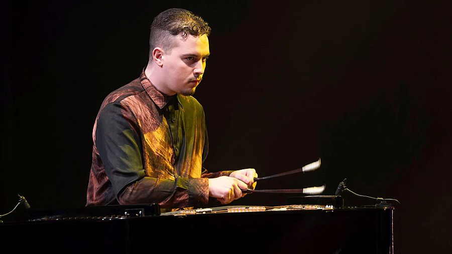 Áron Horváth, músico de percusión, toca un instrumento de láminas con cuatro baquetas. Viste camisa estampada vibrante. Fondo oscuro, iluminación cálida.