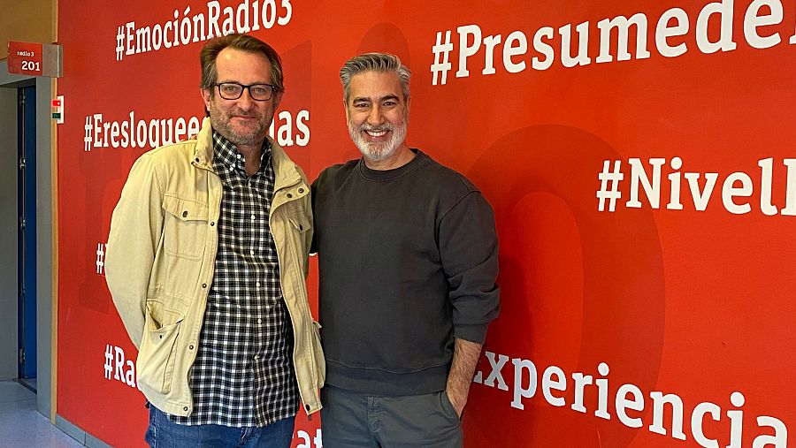 Carlos Castrejón y Arturo Martín sonríen ante una pared roja con hashtags de Radio 3. Carlos lleva gafas y chaqueta beige; Arturo, sudadera gris y apoya su brazo en el hombro de Carlos.