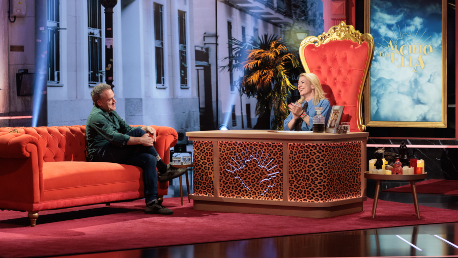 Daniel Guzmán y una presentadora rubia conversan en un plató con decoración de calle nocturna, sofá naranja, sillón trono y un escritorio con estampado de leopardo. El programa se titula 'Al cielo con ella'.