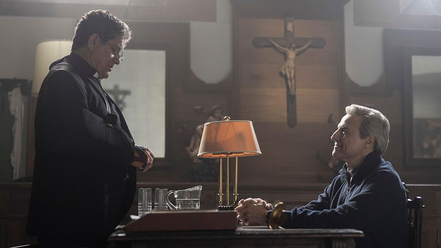 Fotograma de 'La luz': clérigo con cabestrillo y José Coronado conversan en un estudio con crucifijo y lámpara de escritorio.