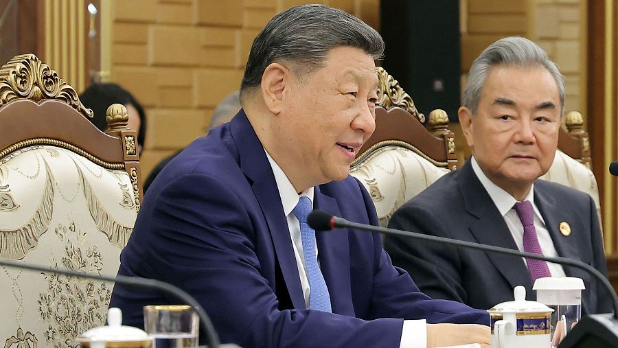 Xi Jinping y Wang Yi, dos figuras asiáticas, participan en una reunión formal. Xi Jinping viste traje azul y sonríe, mientras Wang Yi, con traje oscuro, muestra una expresión seria.