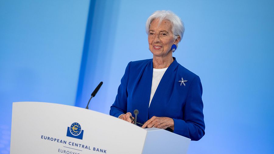 Christine Lagarde, presidenta del BCE, sonríe en una rueda de prensa con blazer azul cobalto y pendientes ovalados. El atril muestra el logo del BCE y dos micrófonos.