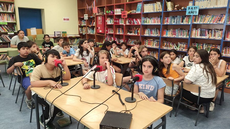 Niños de 9 a 12 años graban un programa de radio en una biblioteca escolar, con micrófonos RNE y un cartel "ON AIR".