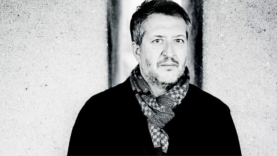 Retrato en blanco y negro de Thomas Adès, compositor de mediana edad, con cabello y barba entrecanos, y una bufanda geométrica.