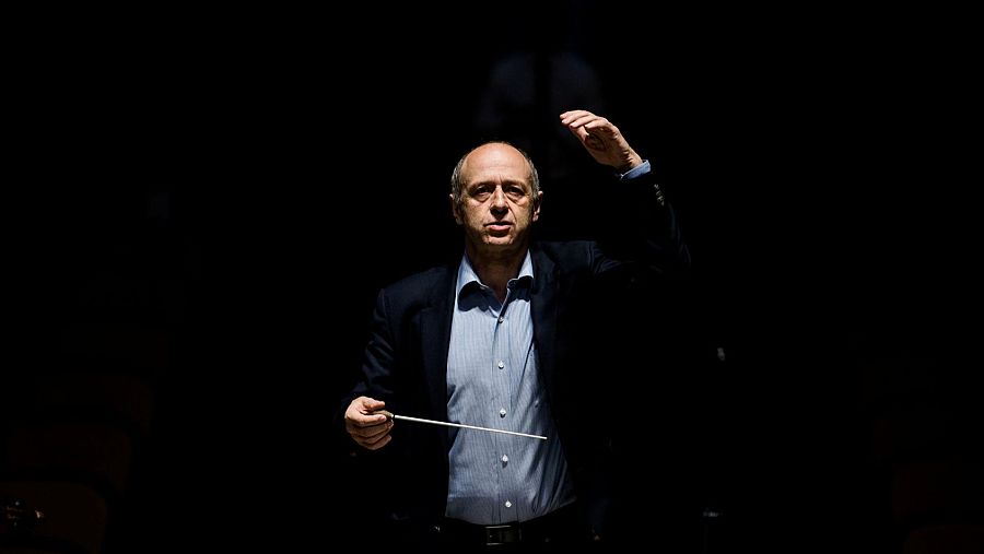 Retrato de Iván Fischer dirigiendo, con chaqueta oscura, camisa azul claro y batuta. Luz frontal intensa sobre fondo negro.