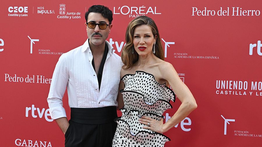 Jaydy Michel y un hombre posan en un photocall rojo con logotipos de patrocinadores. Ella lleva un vestido blanco de lunares negros y él gafas de sol y fajín.