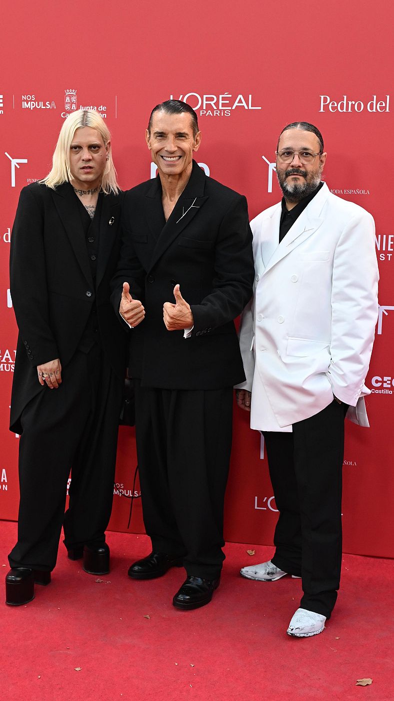 Jesús Vázquez, con traje negro y pulgares arriba, posa con dos diseñadores de Acromatryx en un evento con fondo rojo y logotipos de patrocinadores.