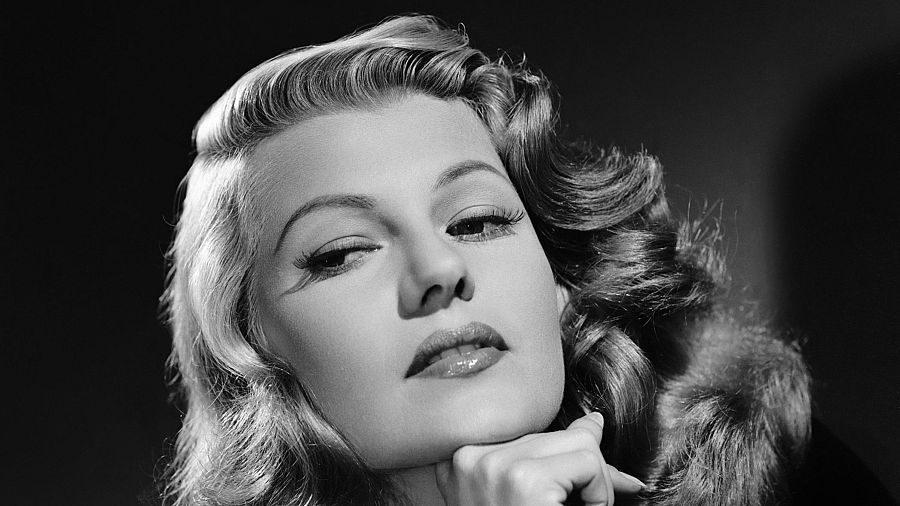 Primer plano en blanco y negro de Rita Hayworth, con cabello ondulado, mirada seductora y mano apoyada en la barbilla. Fondo oscuro.