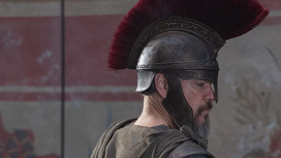 Matt Damon con casco de guerrero antiguo, barba gris y túnica de color tierra. Fondo desenfocado con pared de tonos rojizos y claros.