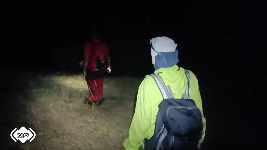 Dos excursionistas, uno con chaqueta amarilla reflectante y otro con ropa roja y bastón, avanzan por un sendero nocturno. Se distingue el logo "sepa emergencias".