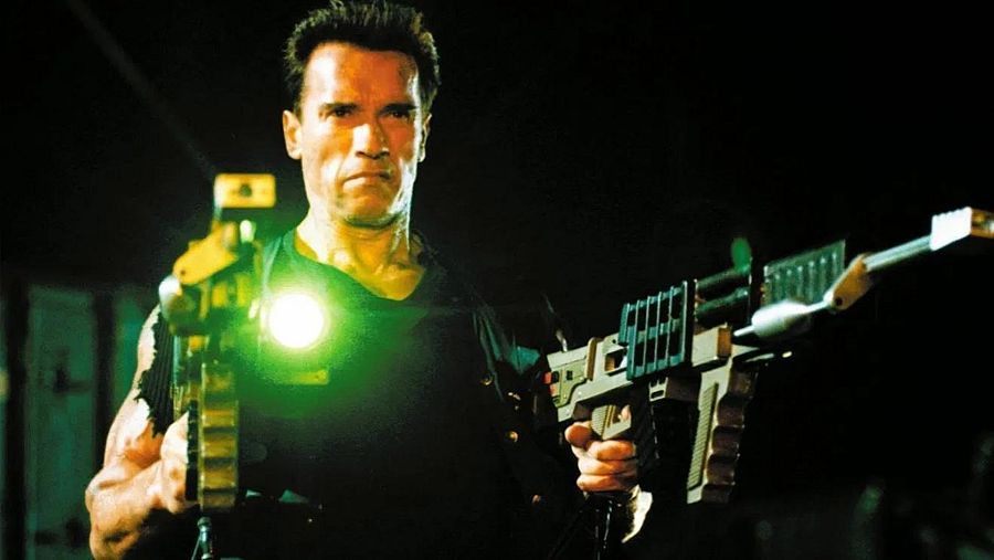 Arnold Schwarzenegger en pose de acción con dos armas futuristas, una de ellas emite una luz verde intensa. Fondo oscuro.