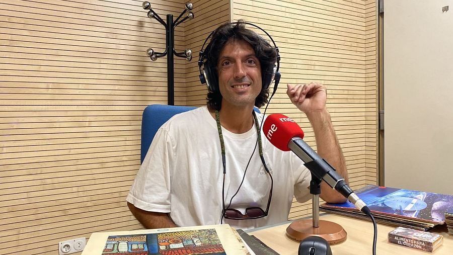 Alejandro Jiménez sonríe en un estudio de radio con auriculares y gafas de sol. Un micrófono "rne" rojo y varios objetos decoran la mesa frente a él.