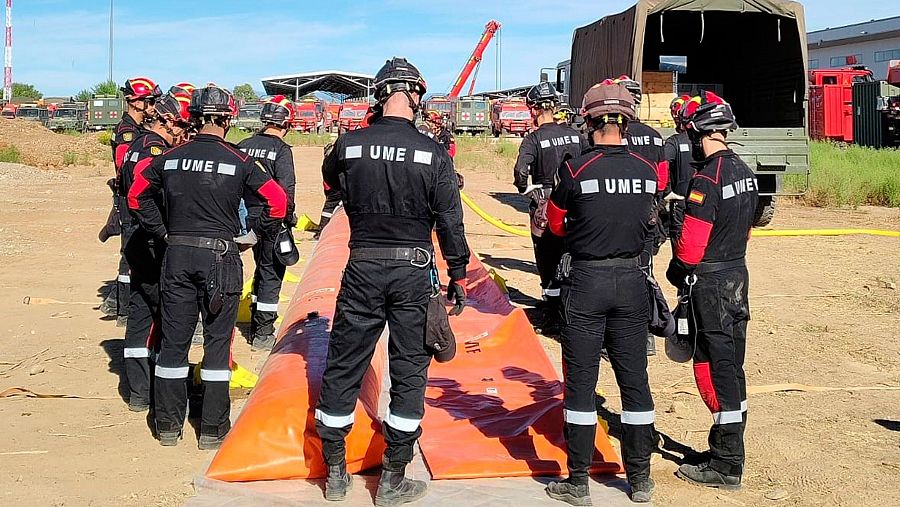 Miembros de la UME, uniformados y equipados, se preparan junto a una gran estructura inflable naranja y mangueras amarillas en un terreno seco, con vehículos de emergencia al fondo.