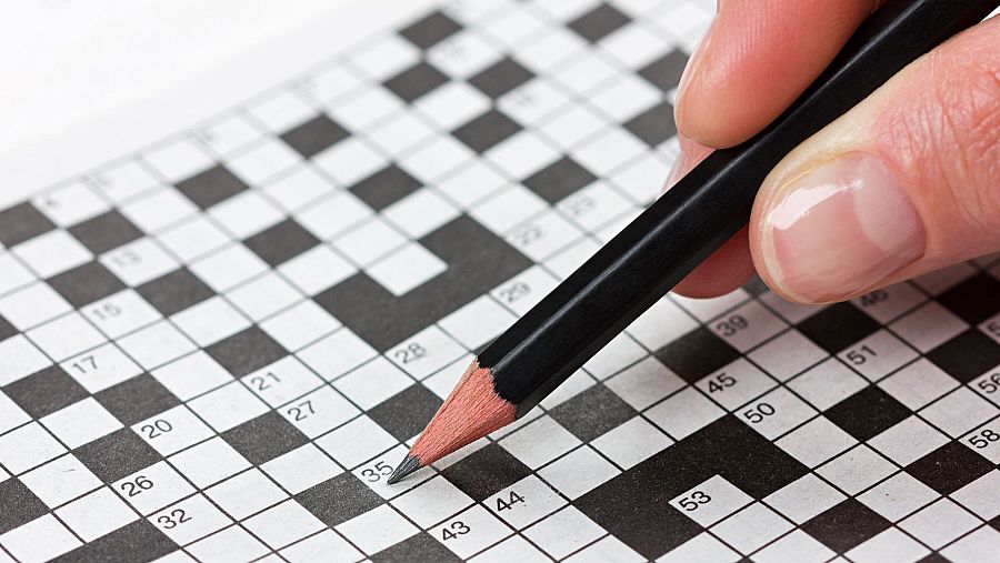 Primer plano de una mano con lápiz sobre un crucigrama, con números visibles como 15, 17, 20, 21, 26, 27, 28, 29, 32, 35, 39, 43, 44, 45, 50, 51, 53, 56 y 58. El lápiz se posiciona sobre el cuadrado 35.