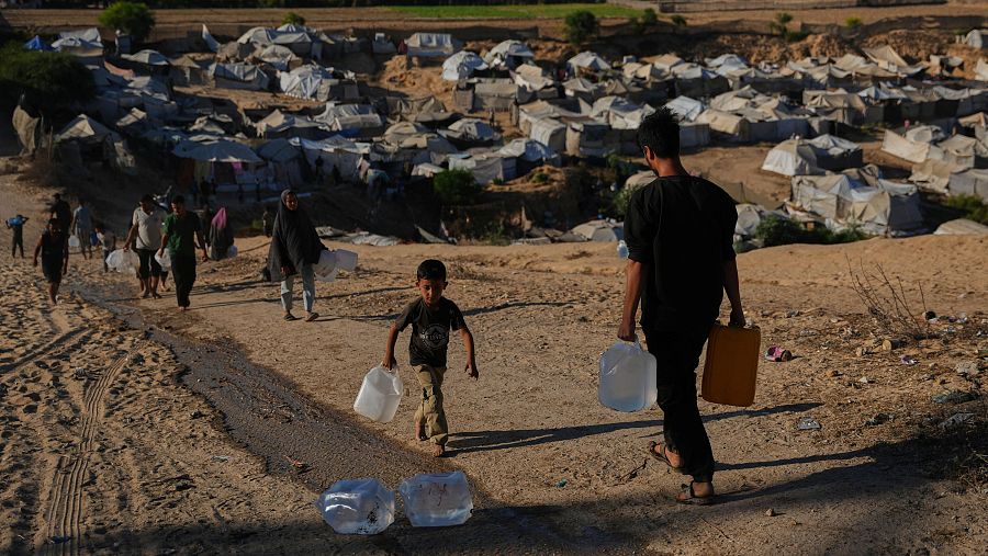 Personas desplazadas transportan garrafas de agua por una colina seca junto a un extenso campamento de refugiados con tiendas improvisadas en Gaza.