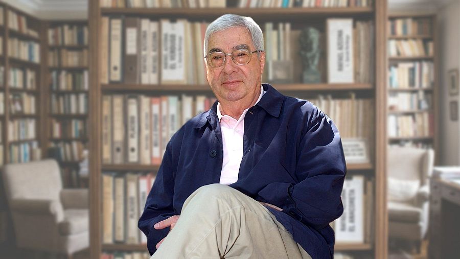 El escritor Rafael Azcona posa sentado en una biblioteca con estanterías de madera, archivadores y libros al fondo.