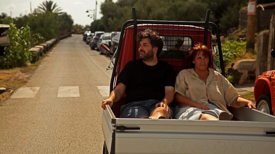 Dos personas viajan sentadas en la parte trasera de un motocarro rojo por una carretera rural rodeada de vegetación bajo una luz solar dorada.