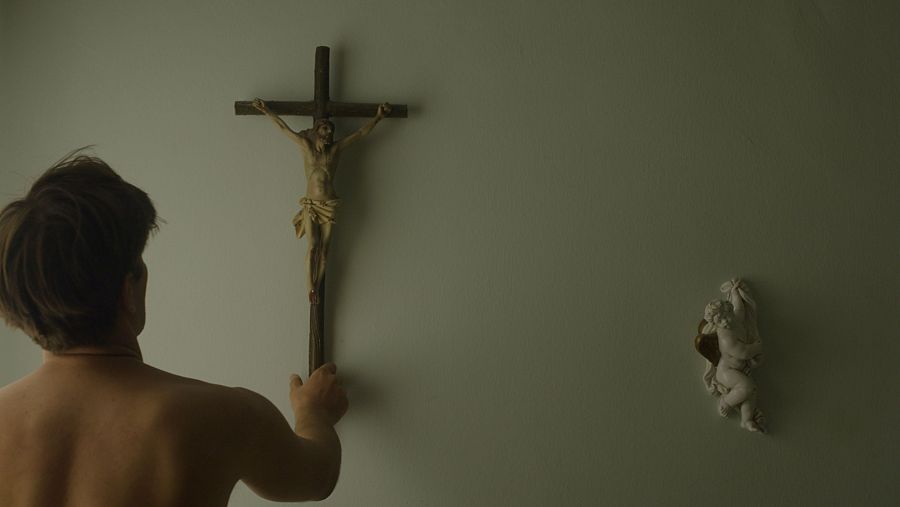 Joven de espaldas con el torso desnudo tocando un crucifijo de madera colgado en una pared con iluminación tenue.