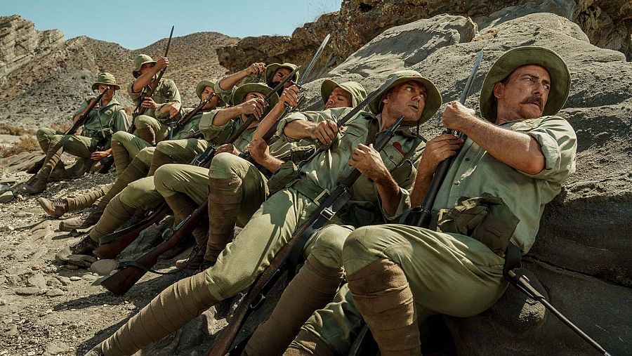 Soldados con uniformes de época colonial y fusiles con bayoneta descansan en alerta sobre una ladera rocosa y árida bajo un cielo despejado.