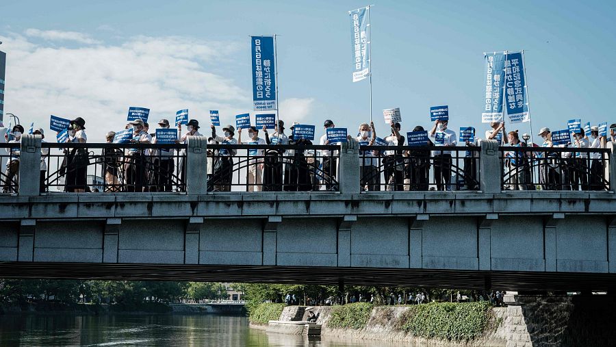 Manifestantes con pancartas que dicen "Oren en silencio por la paz" se paran en un puente sobre el río Motoyasu en Hiroshima el 6 de agosto de 2021.