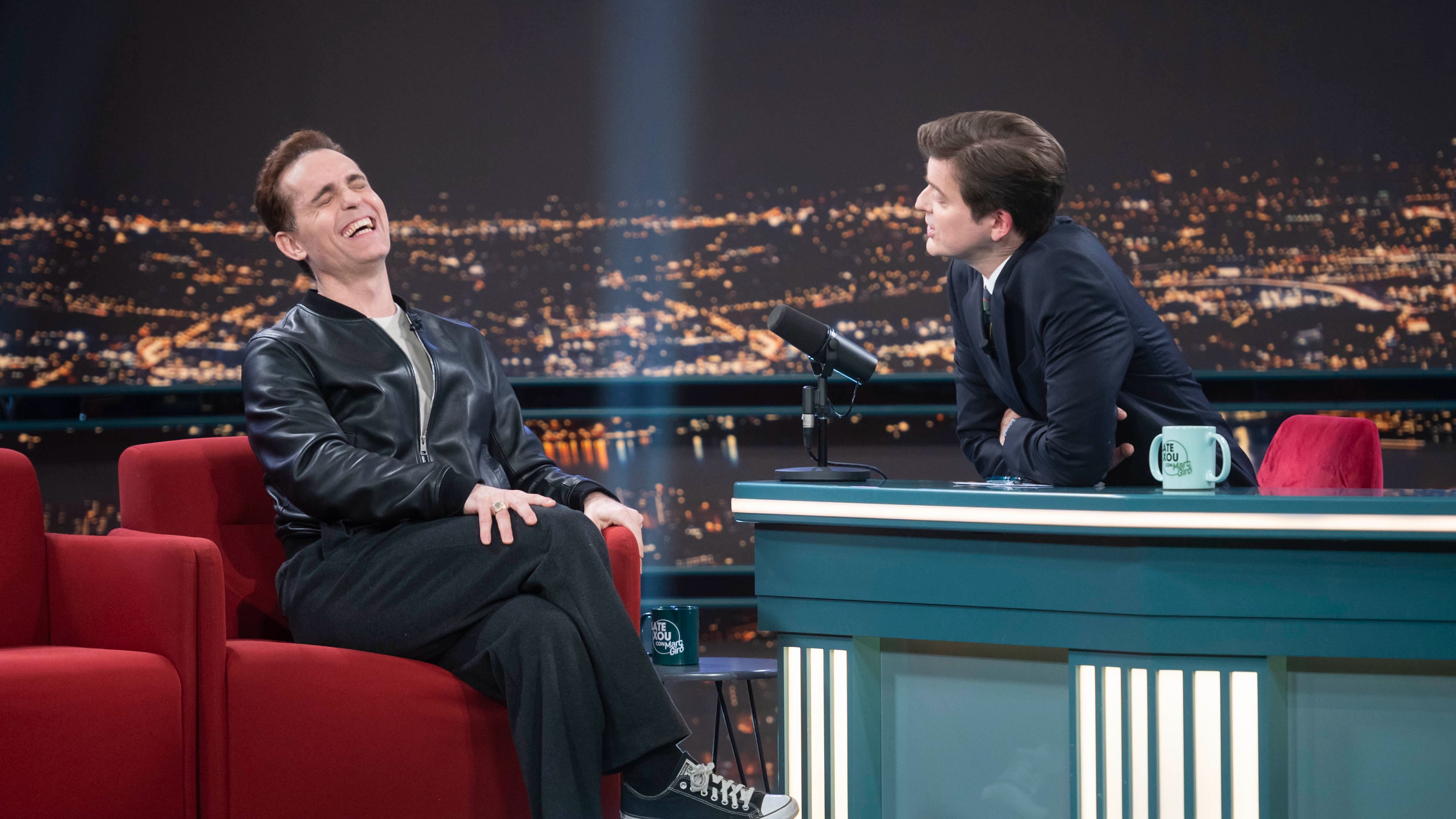 Pedro Alonso, actor conocido por 'La casa de papel', ríe en una entrevista televisiva sentado en un sofá rojo, con chaqueta negra y zapatillas Converse. El presentador le observa desde una mesa cercana.