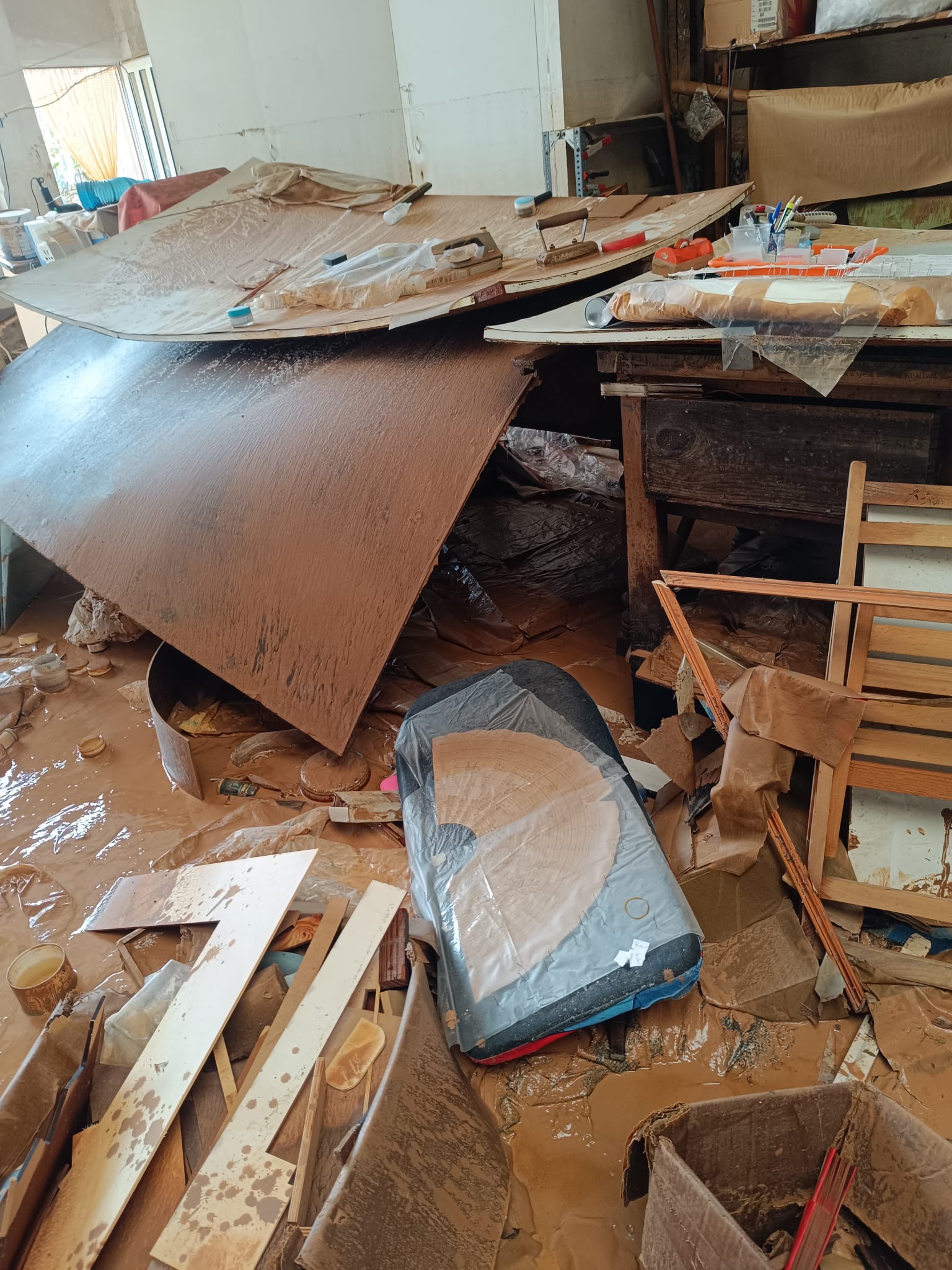 Así quedó el taller de Abanicos Andrés tras la dana del 29 de octubre | Fotos cedidas por Macarena Andrés Un taller de abanicos devastado por una inundación, con agua marrón cubriendo el suelo y escombros esparcidos. Se ven mesas de trabajo con barro, una silla rota y abanicos deteriorados.