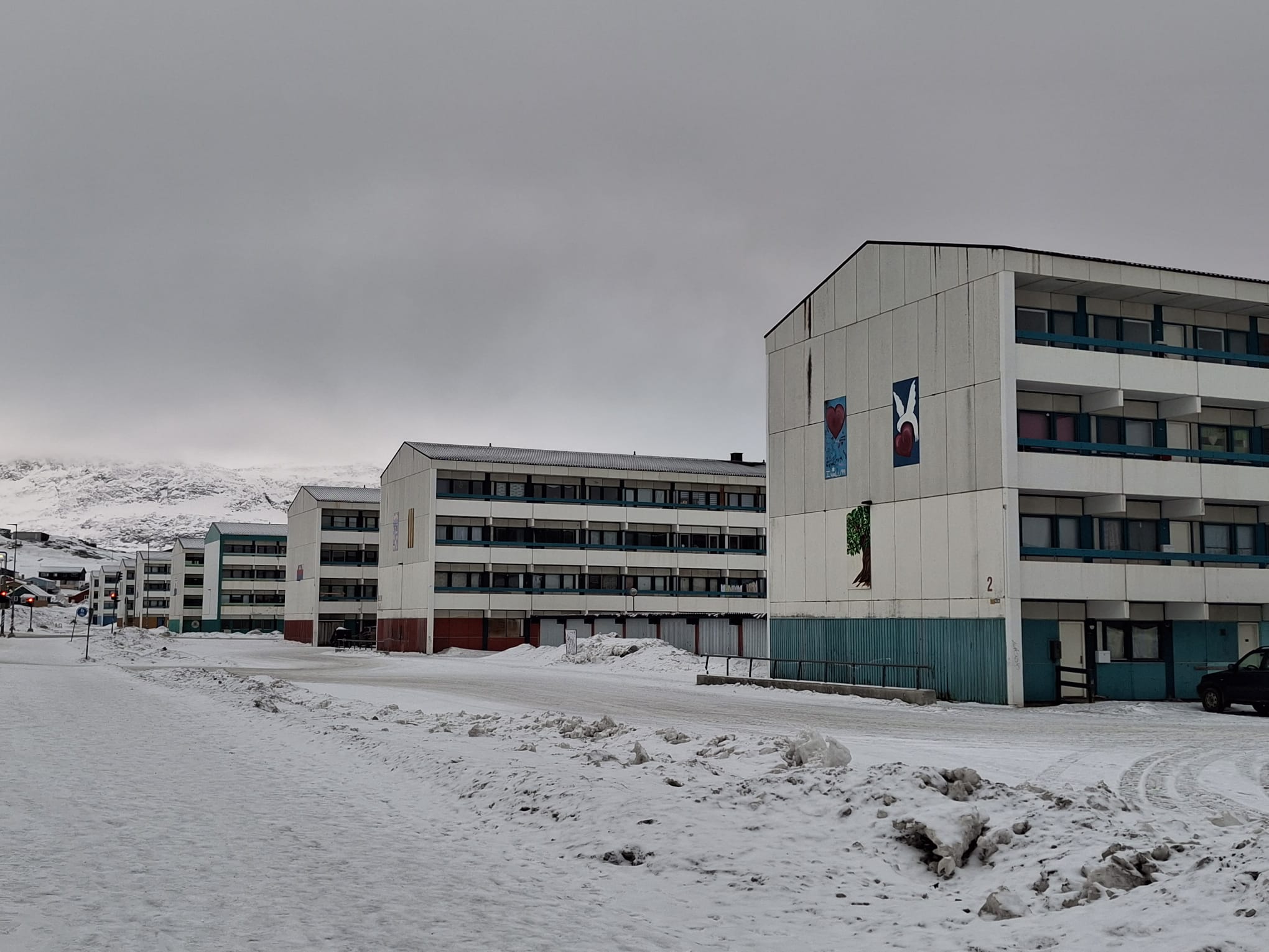 Edificios blancos de varios pisos con detalles verdes y murales en un paisaje nevado. Nieve acumulada y un coche negro visible.