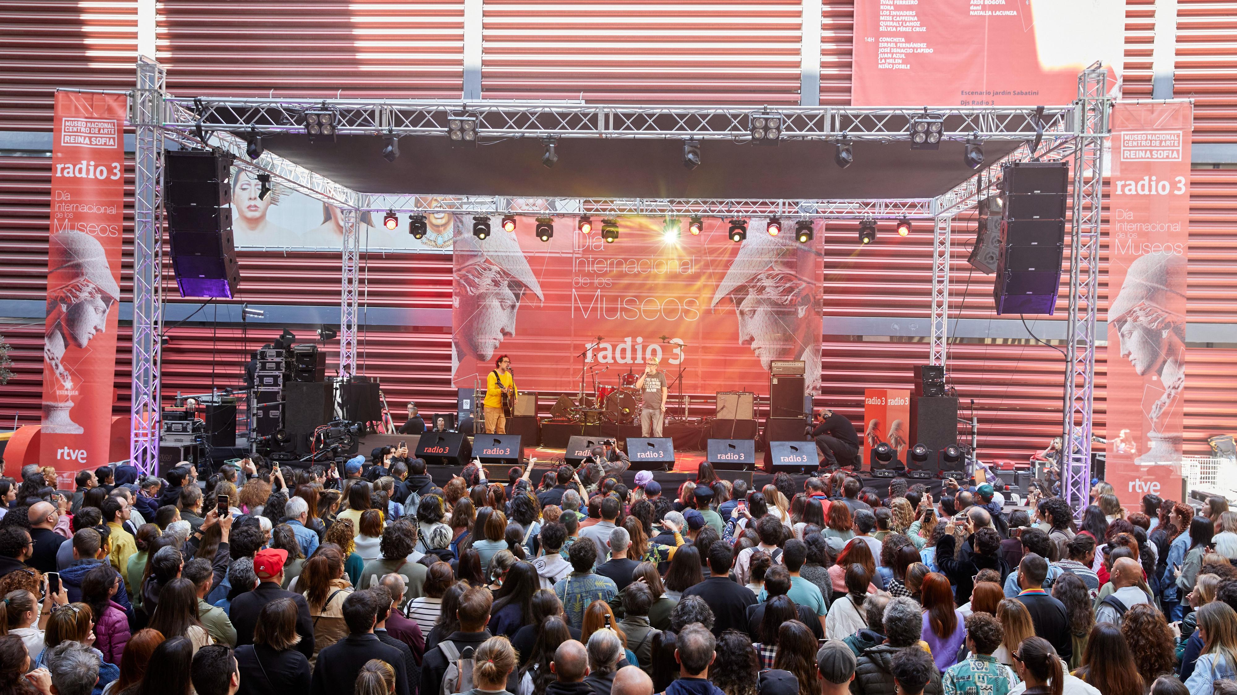 Concierto en exterior con músicos, gran banner "Día Internacional de los Museos" y logo RTVE. Público disfrutando del evento.