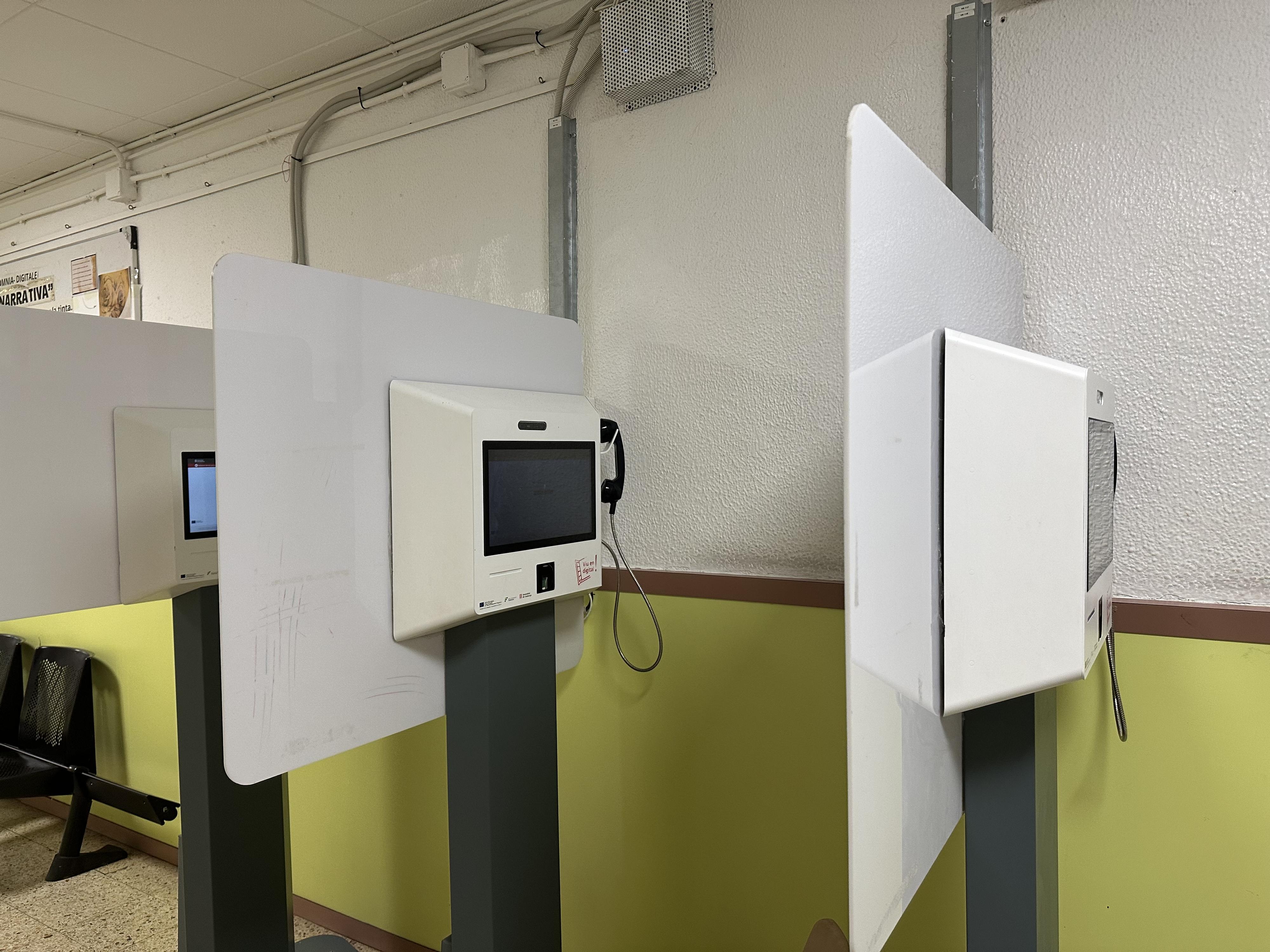 Espacio para hacer videollamadas a familiares Se observan varias cabinas individuales para videollamadas, equipadas con pantallas, teléfonos y paneles protectores blancos. El entorno presenta paredes claras con una sección inferior amarilla y sillas negras en el fondo, sugiriendo un área de espera.