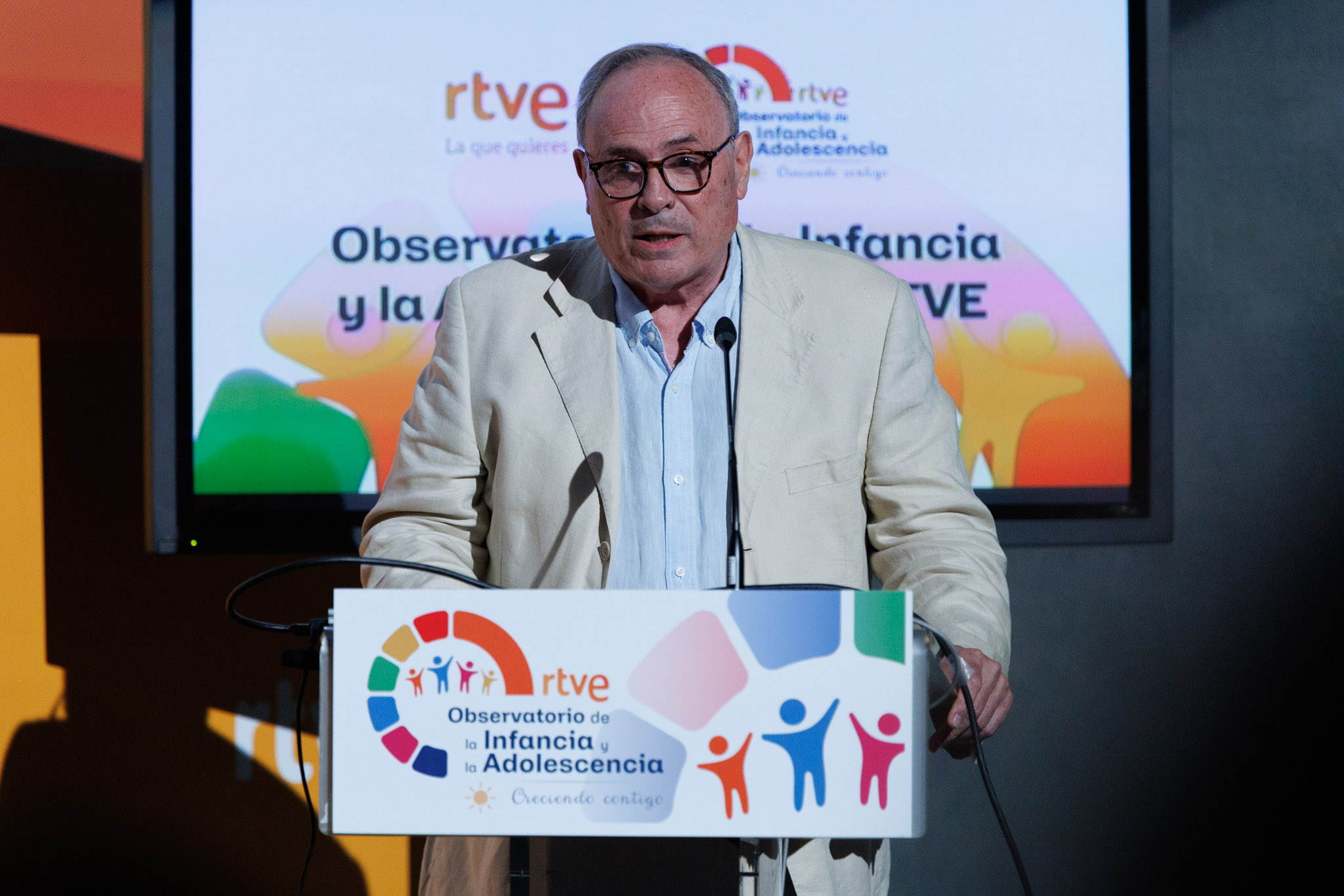 Esteban Mayoral Esteban Mayoral habla detrás de un atril con logos de RTVE y "Observatorio de la Infancia y la Adolescencia", lema "Creciendo contigo", y siluetas de niños y personas en pantalla.