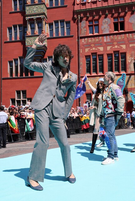 Los 'looks' de la Alfombra Turquesa en Eurovisión 2025 | Australia: Go-Jo Un hombre en traje gris y gafas de sol, y otras personas, caminan por una alfombra turquesa en un evento, con banderas australianas visibles en el fondo.